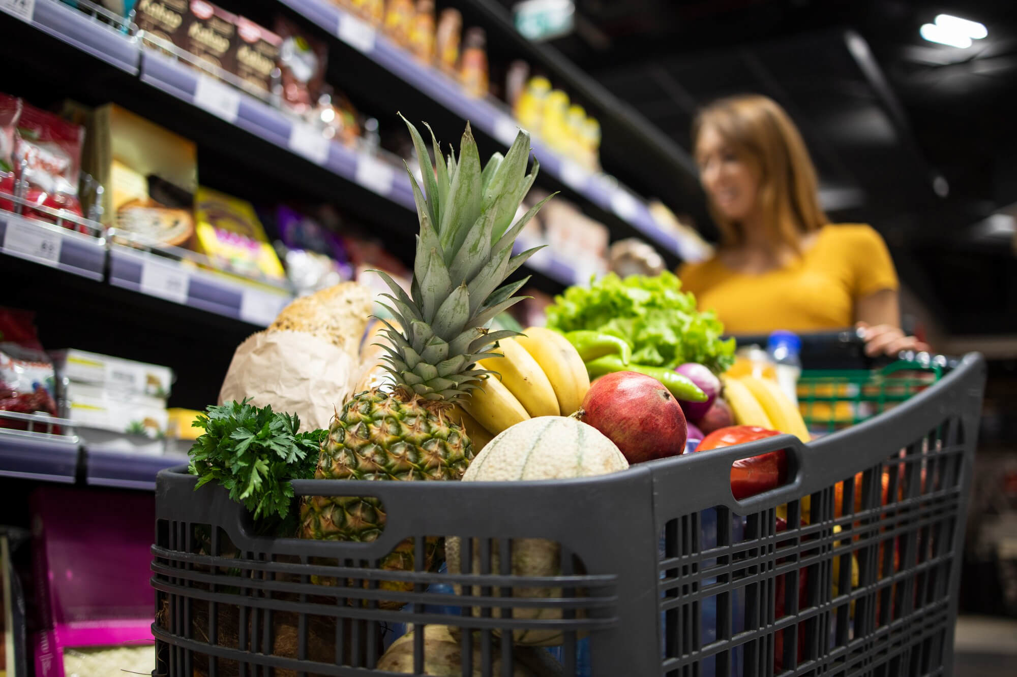 close-up-view-shopping-cart-overloaded-with-food-while-background-female-person-choosing-products